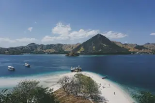 Ilustrasi: Pemandangan pesisir berpasir putih dengan perahu dan perbukitan di latar belakang.