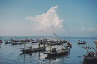 Ilustrasi: Deretan perahu nelayan tradisional bersandar di perairan tenang di bawah awan putih besar.