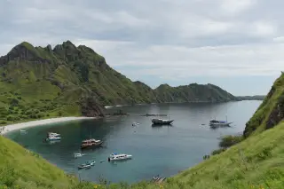 Ilustrasi: Pemandangan teluk berpasir putih dan perbukitan hijau dengan beberapa kapal bersandar di perairan tenang.