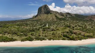 Sebuah mercusuar putih berdiri di pulau berhutan palem dengan gunung besar di latar belakang dan pantai berpasir. Nuca Molas, Mules Island, Flores, Indonesia.,…