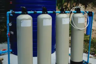 Ilustrasi: Empat tabung filter pengolahan air berwarna krem dengan instalasi pipa biru di luar ruangan.