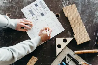 Ilustrasi: Tangan seseorang sedang menggambar sketsa arsitektur di meja kerja kayu dengan penggaris dan perkakas.