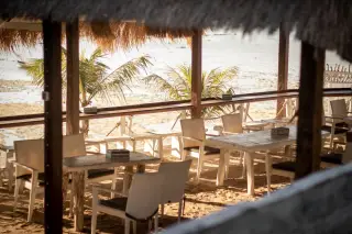 Ilustrasi: Area meja dan kursi restoran beratap jerami di tepi pantai berpasir dengan latar pohon kelapa.