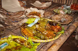 Ilustrasi: Makanan bungkus daun pisang dan piring anyaman bambu di atas meja kayu.