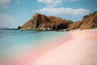 Ilustrasi: Pantai berpasir merah muda dengan air laut tenang dan bukit berbatu di NTT.