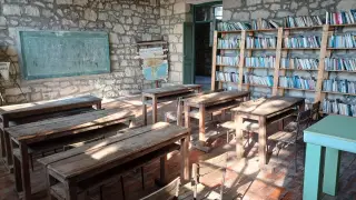 Ilustrasi: Ruang kelas berdinding batu dengan deretan meja kayu kosong dan rak buku.
