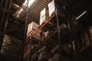 Ilustrasi: Tumpukan kardus dan palet tersusun di rak tinggi di dalam gudang logistik.