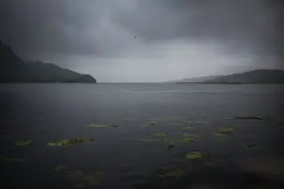 Ilustrasi: Pemandangan perairan tenang berlatar perbukitan di bawah langit mendung gelap.