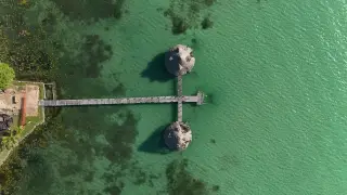 Ilustrasi: Pemandangan udara dermaga kayu dengan dua pondok beratap jerami di atas perairan jernih kehijauan.