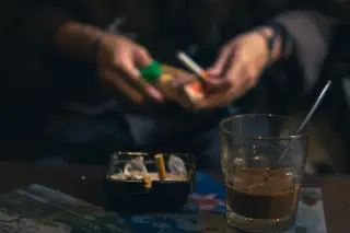 Ilustrasi: Segelas kopi dan asbak berisi puntung rokok di atas meja dengan seseorang memegang rokok di latar belakang.