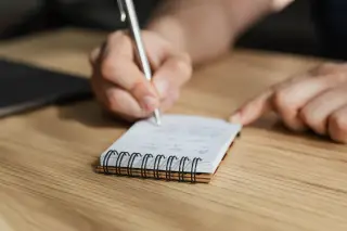 Ilustrasi: Seseorang sedang menulis dengan pena di buku catatan kecil di atas meja kayu.
