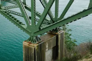 Ilustrasi: Detail struktur rangka jembatan baja berwarna hijau yang berdiri di atas pilar beton di tepi perairan.