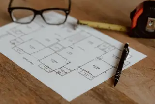 Ilustrasi: Denah rumah, kacamata, meteran gulung, dan pena di atas meja kayu.