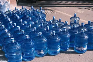 Ilustrasi: Puluhan galon air minum plastik berwarna biru berjejer di atas aspal di bawah sinar matahari.