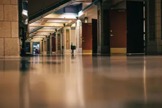 Ilustrasi: Lorong gedung yang sepi dengan lantai mengilap dan deretan pintu kayu terbuka.