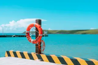 Ilustrasi: Pelampung oranye tergantung di tiang dermaga dengan latar belakang laut tenang dan perbukitan.