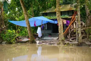Ilustrasi: Tenda terpal biru berdiri di depan rumah seng dekat genangan air keruh.