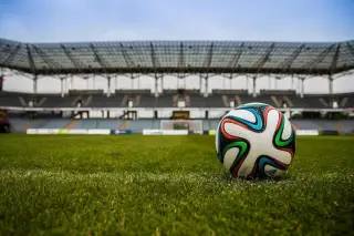 Ilustrasi: Bola sepak bermotif warna-warni tergeletak di atas rumput lapangan dengan latar tribun stadion.