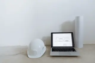 Ilustrasi: Helm proyek putih, laptop menampilkan denah arsitektur, dan gulungan kertas di lantai.
