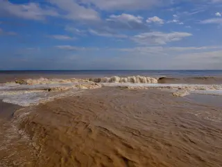 Ilustrasi: Gelombang air laut keruh kecokelatan menerjang pesisir pantai di bawah langit berawan.