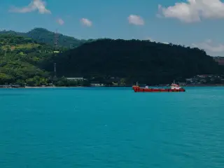 Ilustrasi: Sebuah kapal merah berlayar di perairan laut biru berlatar perbukitan hijau.