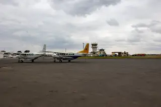Ilustrasi: Dua pesawat perintis terparkir di apron bandara dengan menara kontrol di kejauhan.