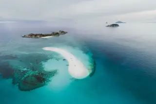 Ilustrasi: Pemandangan udara gosong pasir putih di tengah laut biru jernih dengan pulau-pulau kecil di kejauhan.