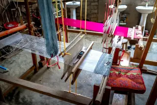 Ilustrasi: Alat tenun tradisional kayu dengan benang tenun dan bantal bermotif di dalam ruangan.