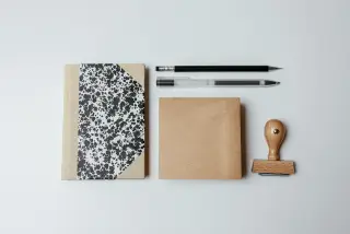 Ilustrasi: Buku catatan, pensil, pena, dan stempel kayu tersusun rapi di atas latar putih.