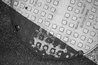 Ilustrasi: Tutup manhole berpola kotak-kotak di atas aspal jalanan secara monokrom.