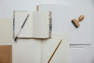 Ilustrasi: Buku catatan terbuka, pulpen, dan sebuah stempel kayu di atas latar putih.
