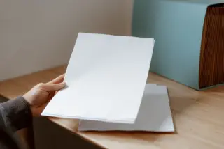 Ilustrasi: Tangan seseorang memegang lembaran kertas putih kosong di atas meja kayu dekat map berkas.