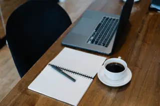 Ilustrasi: Buku catatan polos, pulpen, secangkir kopi, dan laptop di atas meja kayu.