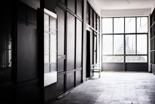 Ilustrasi: Lorong bangunan tua yang kosong dengan pintu kayu dan jendela kaca besar di ujungnya.