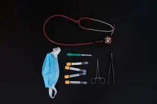 Ilustrasi: Stetoskop, masker medis, jarum suntik, dan tabung sampel tertata di atas latar belakang hitam.
