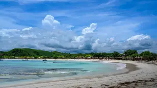 Ilustrasi: Pemandangan pantai berpasir putih dengan air laut jernih dan perbukitan hijau di bawah langit berawan.