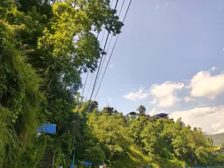 Ilustrasi: Kabel listrik melintang di atas perbukitan hijau dengan rumah-rumah di kejauhan dan langit cerah.