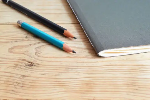 Ilustrasi: Buku catatan dan dua buah pensil tergeletak di atas meja kayu.
