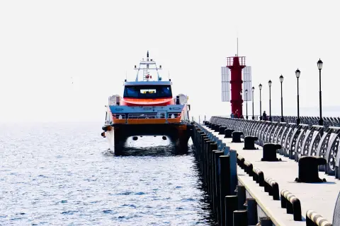 Ilustrasi: Sebuah kapal feri cepat bersandar di samping dermaga panjang dekat menara suar merah.