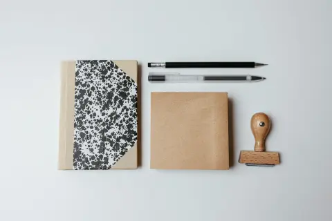 Ilustrasi: Buku catatan, pensil, pena, dan stempel kayu tersusun rapi di atas latar putih.