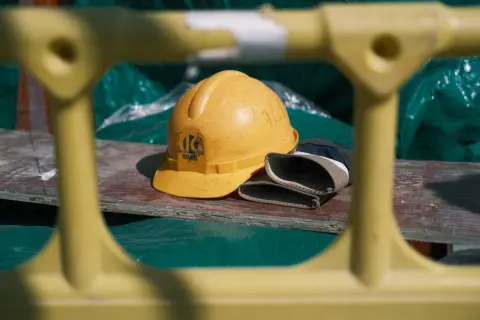 Ilustrasi: Helm proyek kuning dan sarung tangan kerja tergeletak di atas papan kayu.