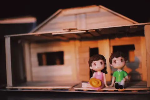 Ilustrasi: Dua figurin kecil duduk di depan miniatur rumah kayu sederhana.