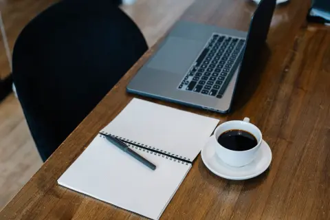 Ilustrasi: Buku catatan polos, pulpen, secangkir kopi, dan laptop di atas meja kayu.