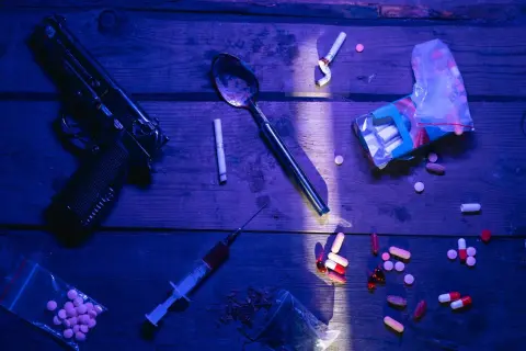 Ilustrasi: Pistol, jarum suntik, sendok, rokok, dan berbagai pil di atas meja kayu dengan pencahayaan dramatis.