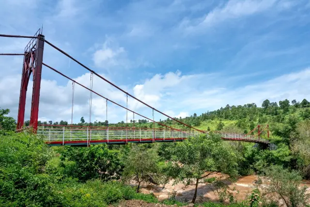 Ilustrasi: Jembatan gantung berkerangka merah melintasi sungai berarus deras di area perbukitan hijau.