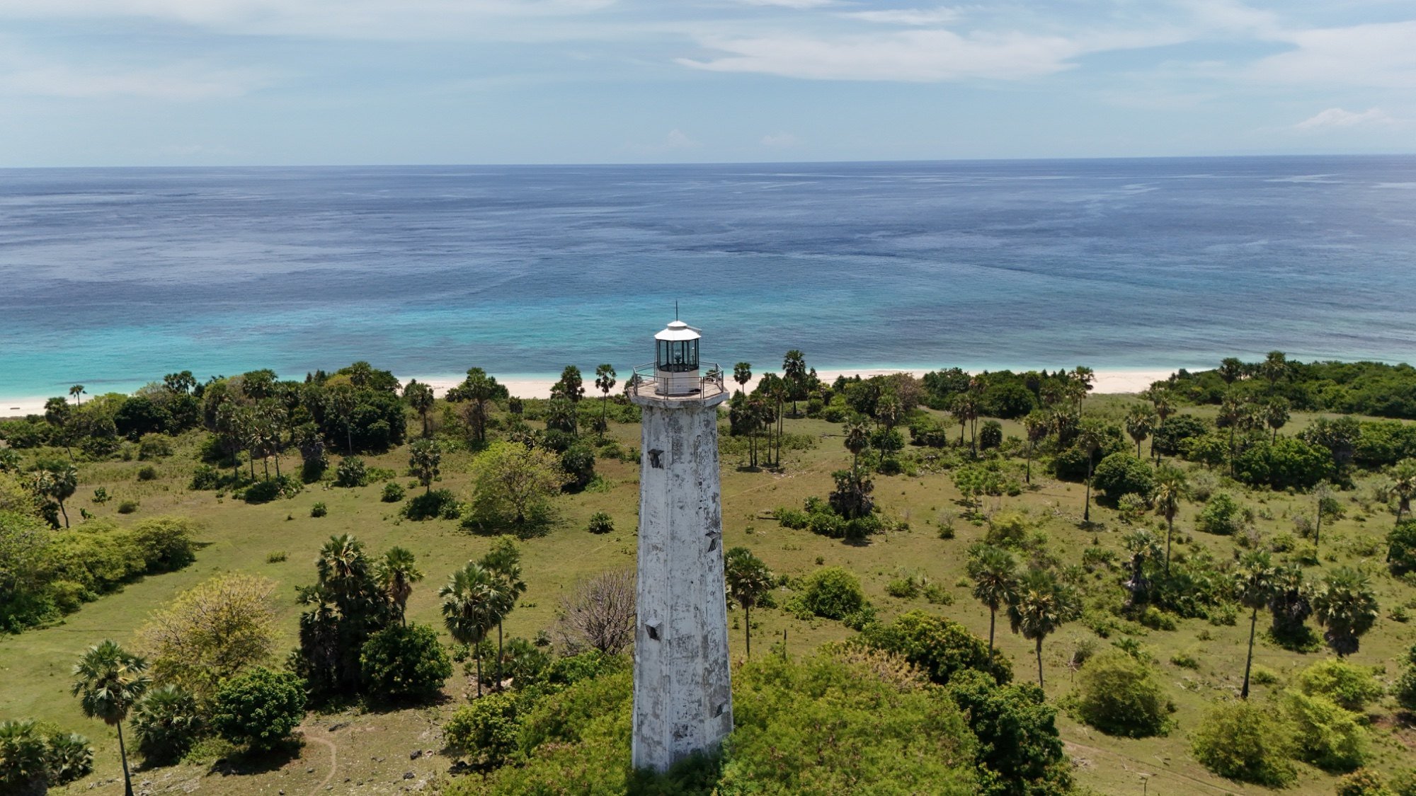 Sebuah mercusuar putih tinggi dikelilingi vegetasi hijau, dengan pantai berpasir dan laut biru di latar belakang. Nuca Molas, Mules Island, Flores, In