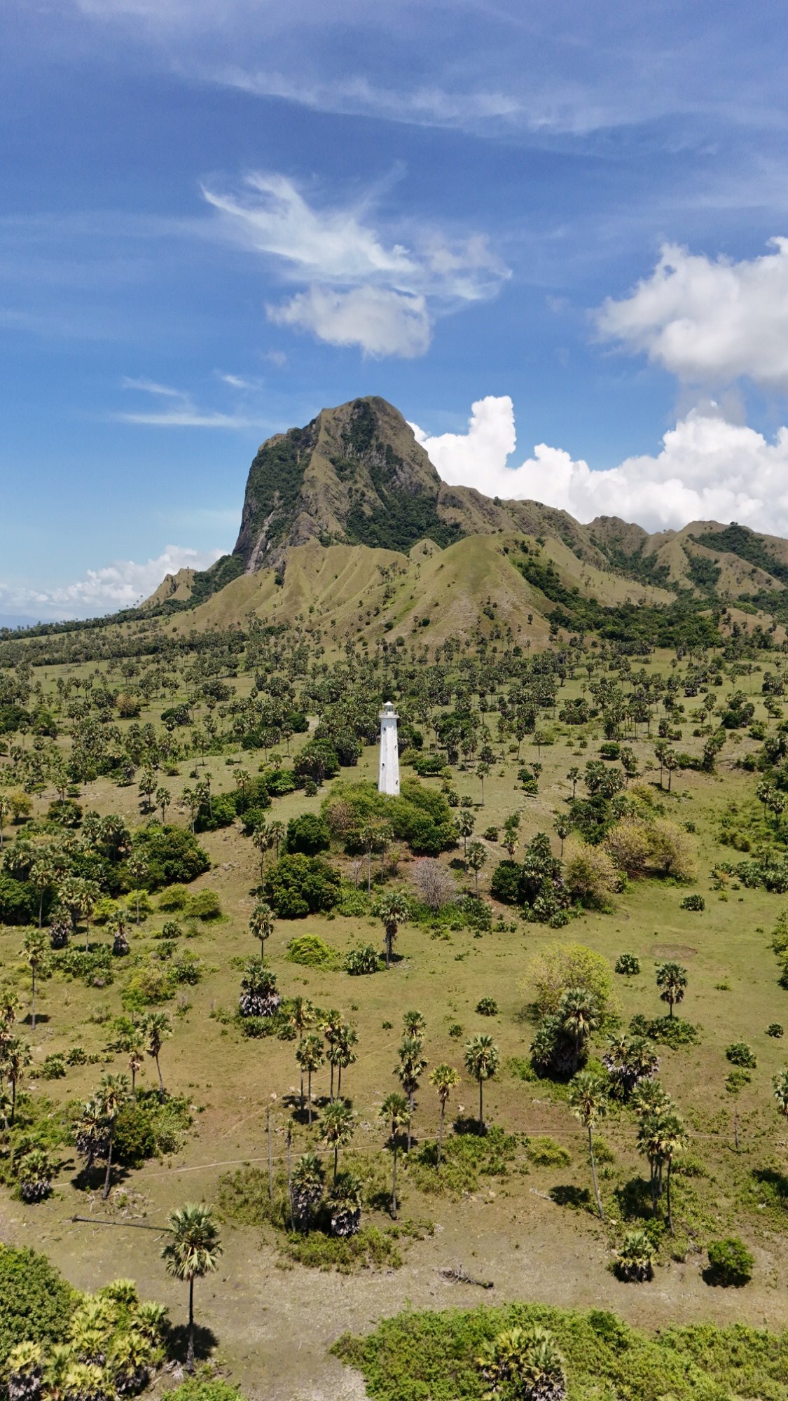 Sebuah mercusuar putih berdiri di tengah pulau bervegetasi hijau dengan gunung besar di latar belakang. Nuca Molas, Mules Island, Flores, Indonesia.,
