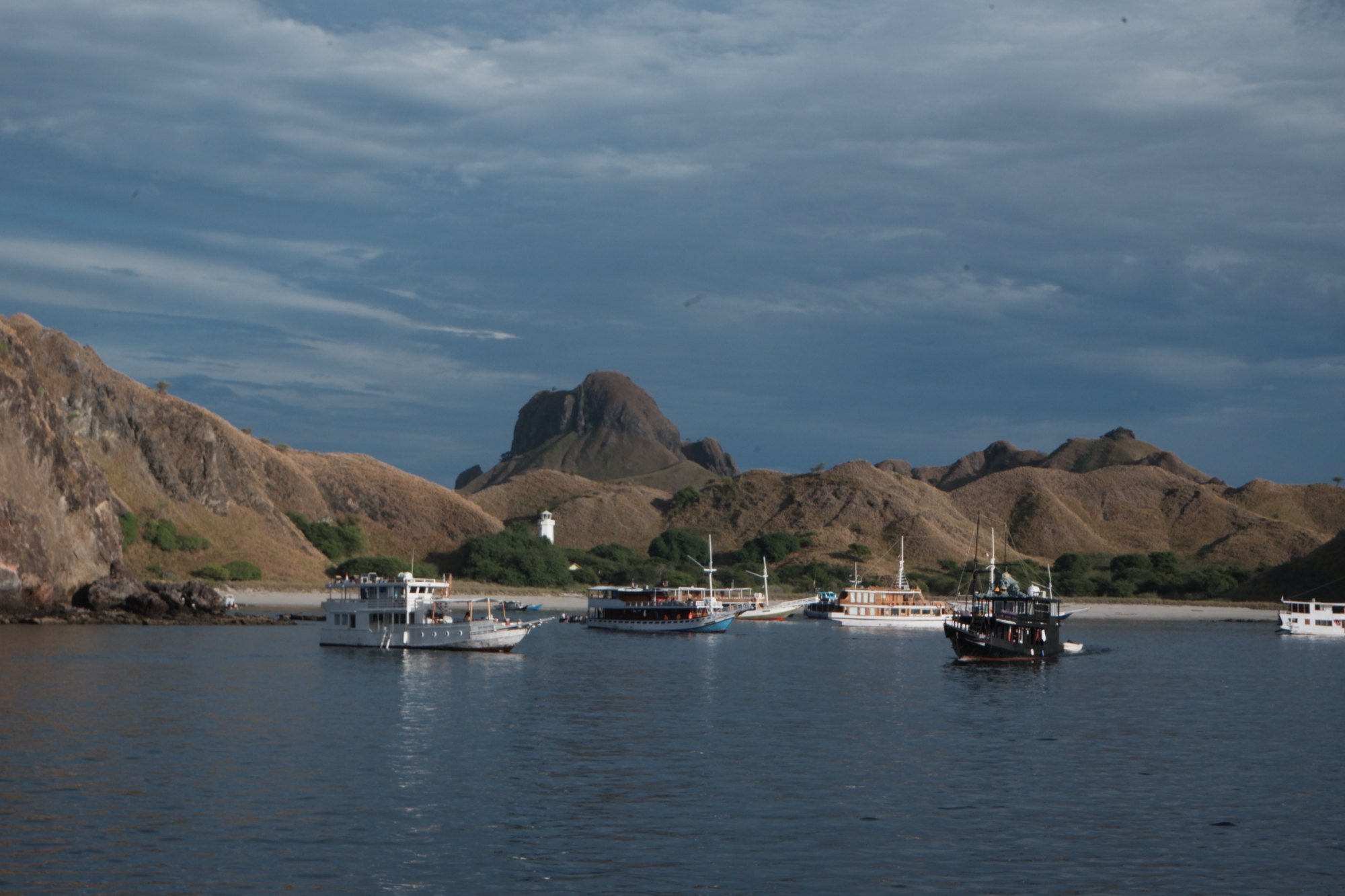 Beberapa perahu berlabuh di teluk dengan latar belakang perbukitan kering dan mercusuar putih di pantai. Pulau Padar, Selasa (22/9/2026).